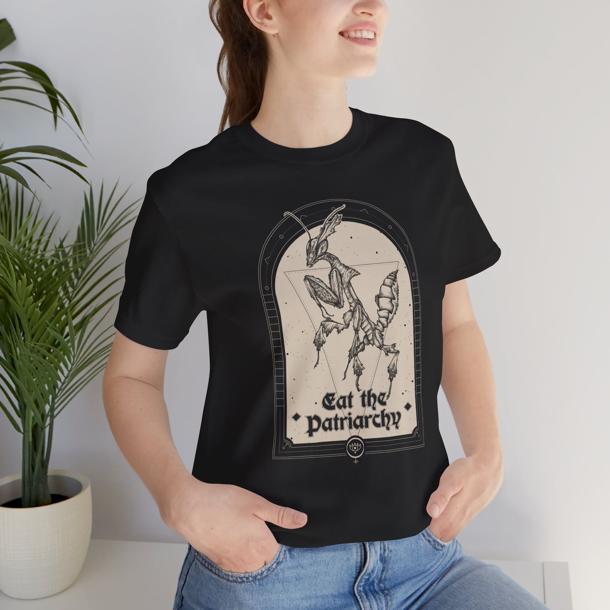 a pretty young adult woman wearing a goth black thirt with a witchy feminist praying mantis illustration that says "eat the patriarchy"