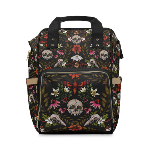 A hauntingly beautiful dark cottagecore multi-functional diaper backpack featuring a goth floral skull pattern in olive green, rust orange, beige, and pink with multiple pockets for the witchy, gothic mom. 