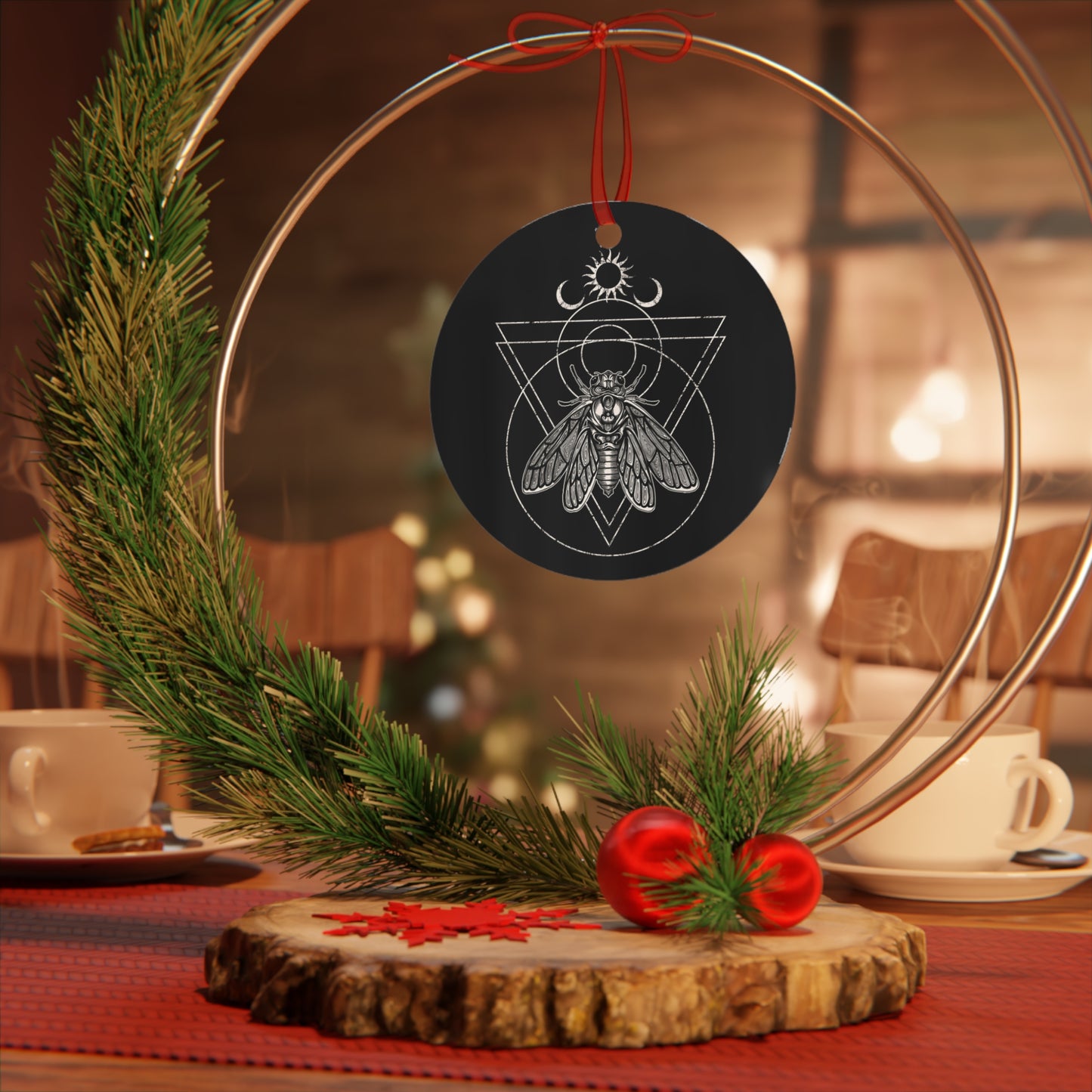 A black aluminum holiday yule tree ornament with a white illustration of a cicada and occult symbols on it, hanging from a red ribbon.
