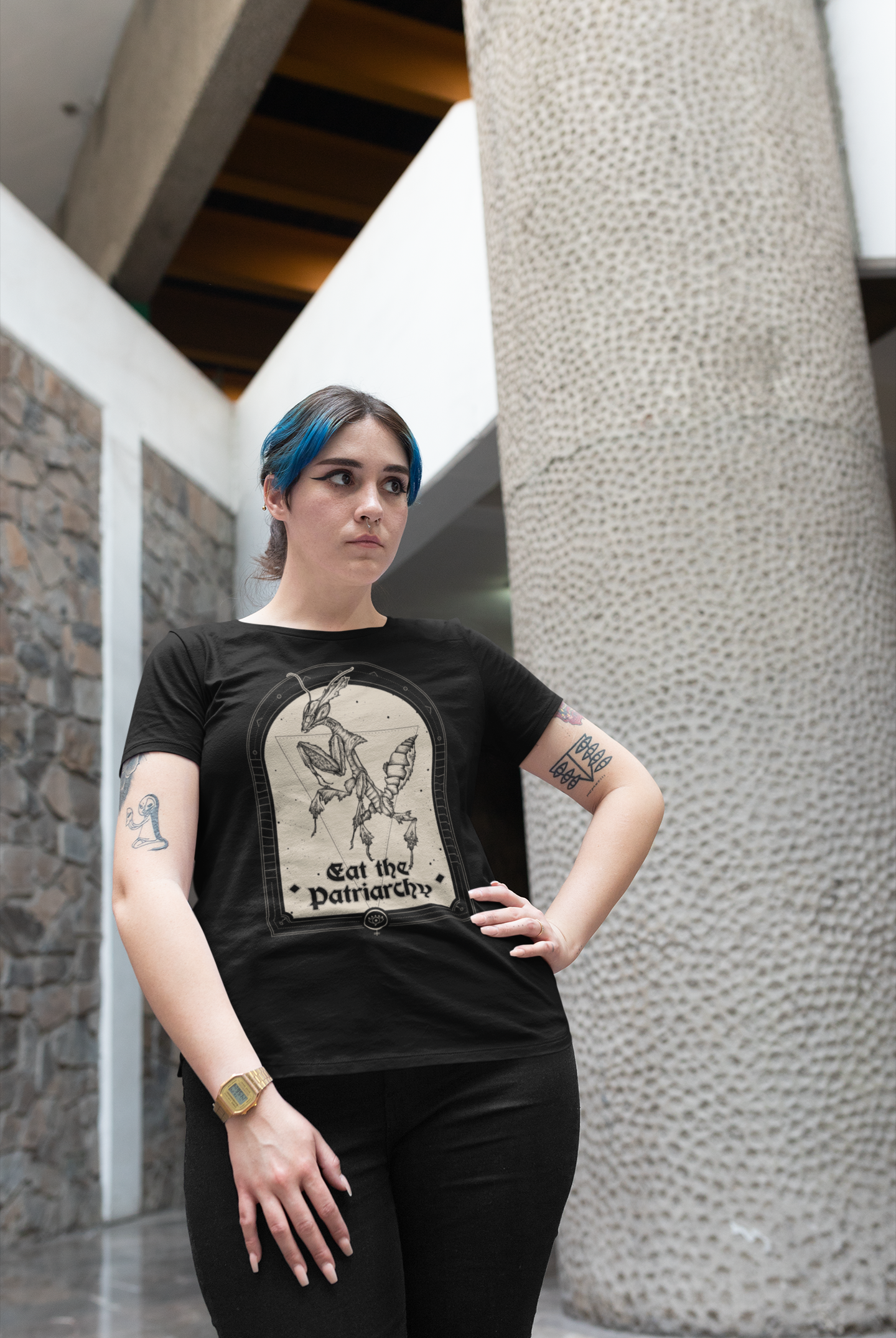 a tattoed young adult woman with blue hair and black cat-eye eyeliner wearing a goth black thirt with a witchy feminist praying mantis illustration that says "eat the patriarchy"