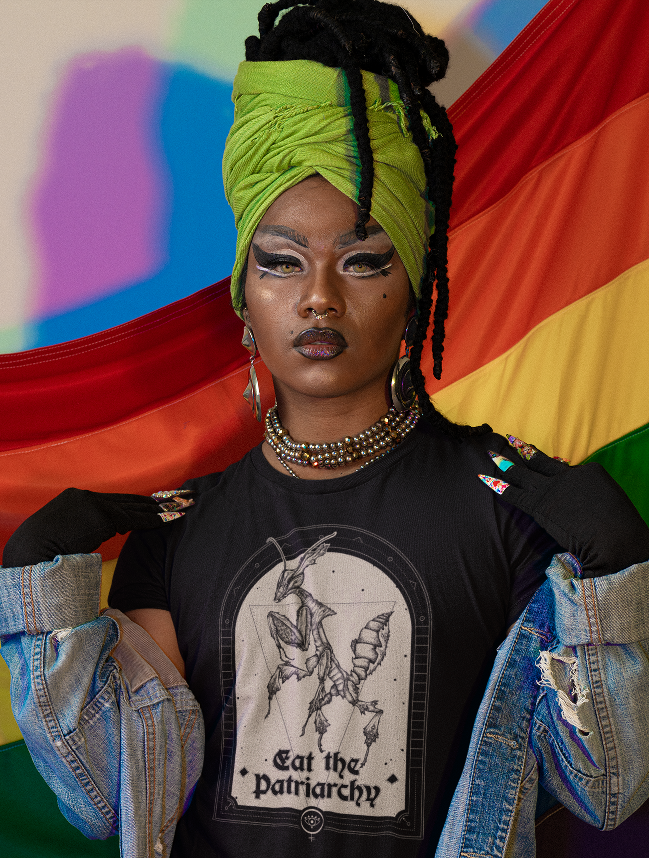 a fashionable black trans woman wearing a green head wrap, punk rock denim jacket, striking makeup, and goth black tee with a witchy feminist praying mantis illustration that says "eat the patriarchy" against a rainbow pride flag