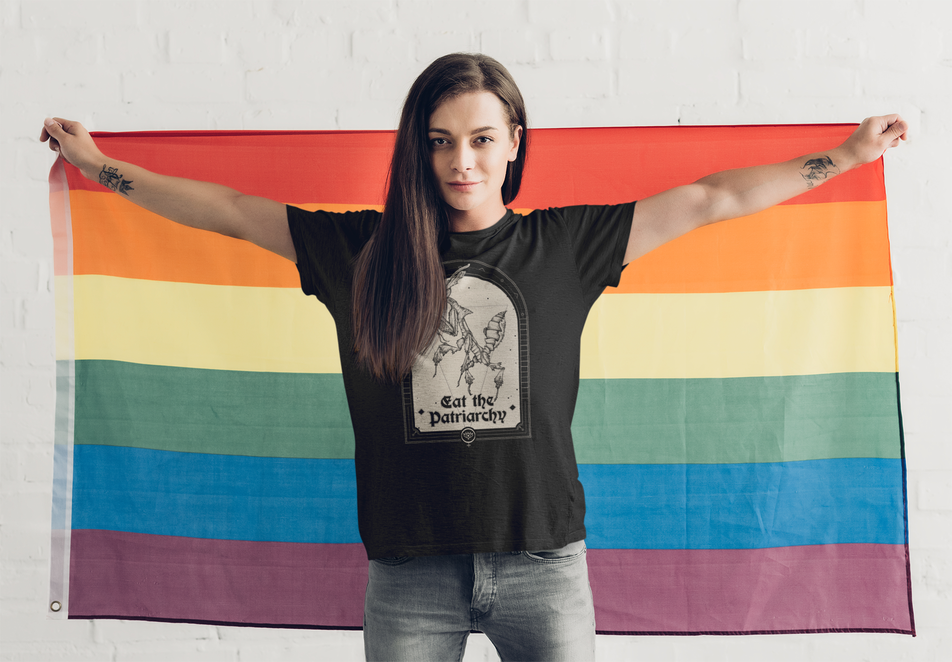 an inclusive activist adult woman with tattoos and brown hair wearing a gothic black top with a witchy feminist praying mantis illustration that says "eat the patriarchy" while holding a rainbow gay pride flag
