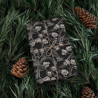 goth Gift wrapped in black floral and skull pattern wrapping paper with brown string, surrounded by green pine needles and pine cones.