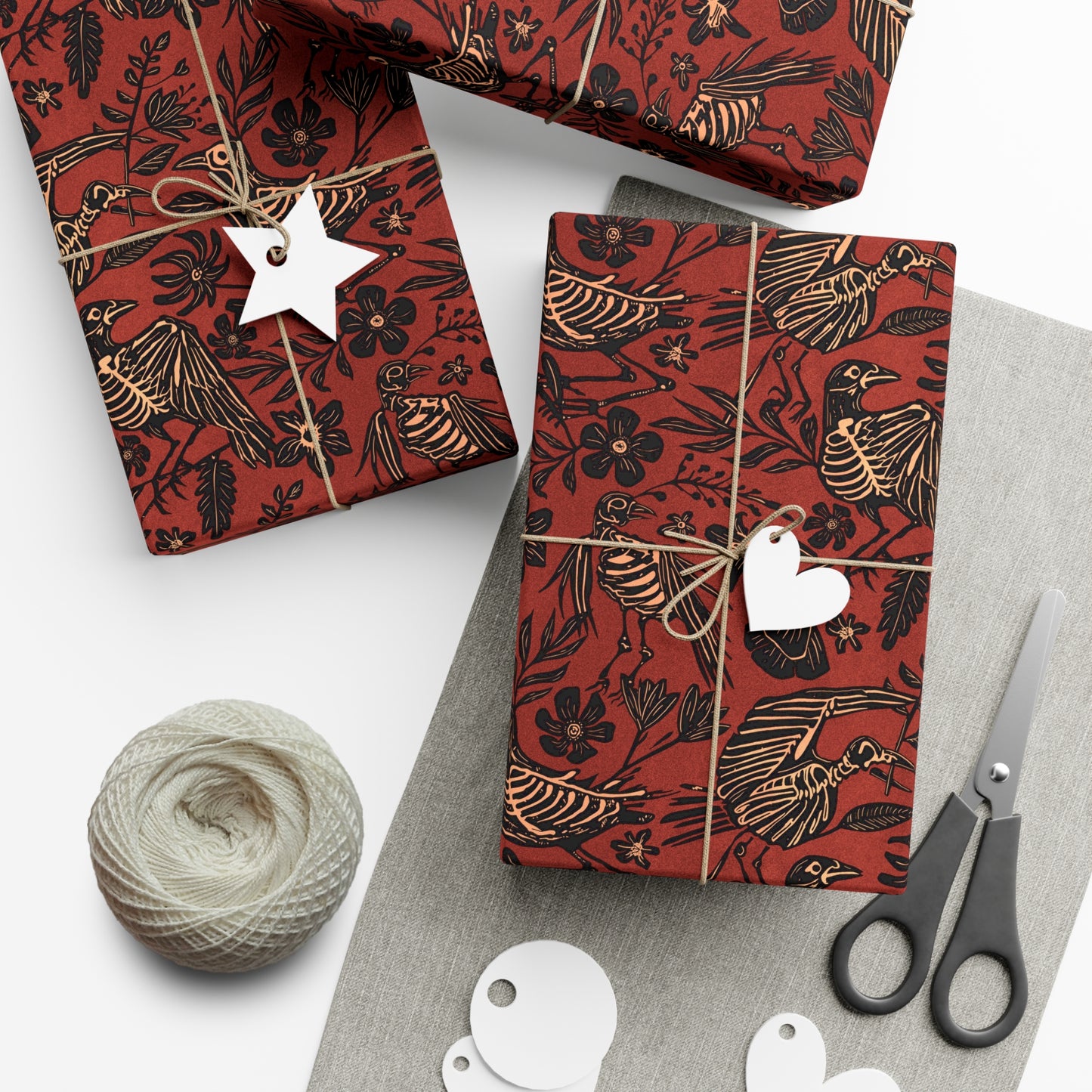 Red gift wrap with black skeleton bird design, scissors, yarn, and white tags on a white surface.