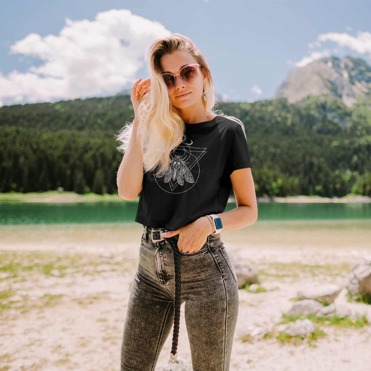 Woman wearing a black gothic cicada t-shirt with a graphic design and sunglasses, standing in a scenic outdoor setting with mountains and a lake.