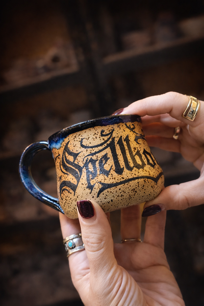 Hand holding a mug with 'Spells' written on it against a blurred background